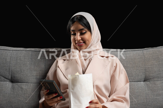 قضاء أوقات ممتعة بمشاهدة الأفلام والمسلسلات، استخدام الأجهزة التكنولوجية الحديثة، بورتريه مقرب لشابة عربية سعودية خليجية مبتسمة ترتدي الحجاب تجلس على كنبة تحمل بيدها كيس الفوشار والهاتف المحمول، ايماءات الاندماج والسرور، خلفية سوداء