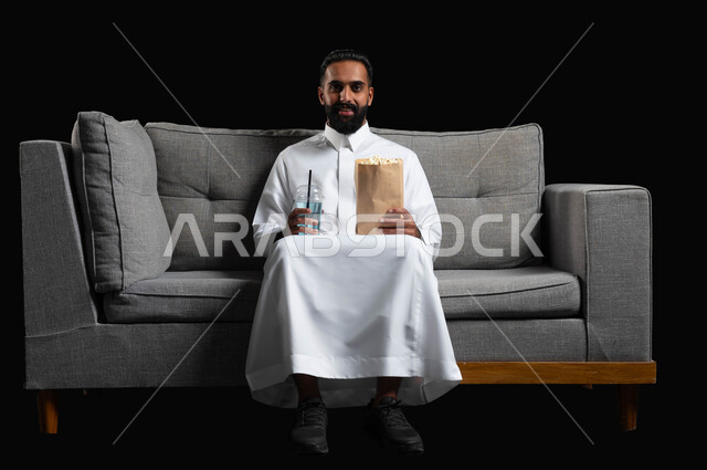 تناول الفوشار والمشروبات الباردة والاستمتاع بمشاهدة الأفلام والمسلسلات، بورتريه لرجل عربي خليجي سعودي مبتسم يرتدي الثوب التقليدي يجلس على اريكة رمادية يقضي أوقات ممتعة ومسلية في المنزل، ايماءات وتعابير الاندماج والسرور، خلفية سوداء