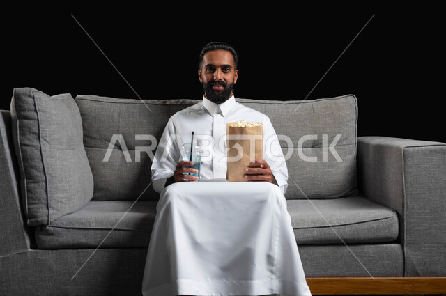 تناول الفوشار والمشروبات الباردة والاستمتاع بمشاهدة الأفلام والمسلسلات، بورتريه لرجل عربي خليجي سعودي مبتسم يرتدي الثوب التقليدي يجلس على اريكة رمادية يقضي أوقات ممتعة ومسلية في المنزل، ايماءات وتعابير الاندماج والسرور، خلفية سوداء