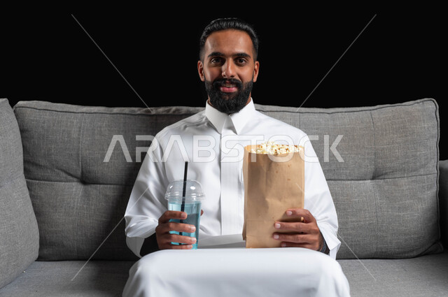 قضاء أوقات ممتعة ومسلية في المنزل، الاستمتاع بمشاهدة الأفلام والمسلسلات، بورتريه مقرب لرجل عربي خليجي سعودي مبتسم يرتدي الثوب التقليدي يجلس على اريكة رمادية يقوم بتناول الفوشار والمشروبات الباردة، ايماءات وتعابير الاندماج والسرور، خلفية سوداء