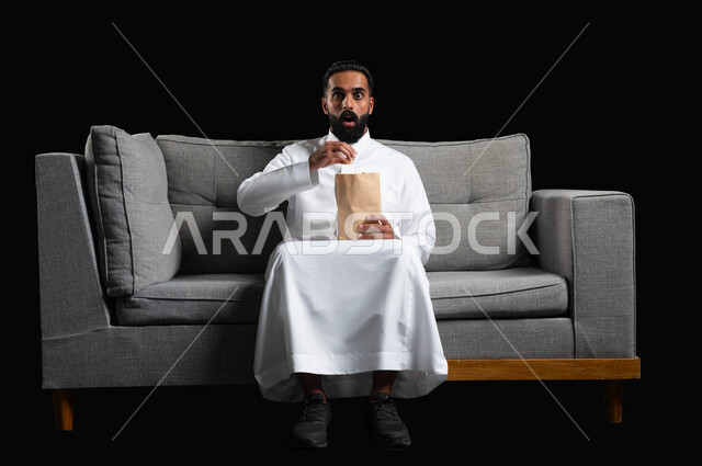 ايماءات وتعابير التعجب والصدمة، الاستمتاع بمشاهدة الأفلام والمسلسلات، بورتريه لرجل عربي خليجي سعودي يرتدي الثوب التقليدي يجلس على اريكة رمادية يقوم بمشاهدة التلفاز وتناول الفوشار،  قضاء أوقات ممتعة ومسلية في المنزل، خلفية سوداء