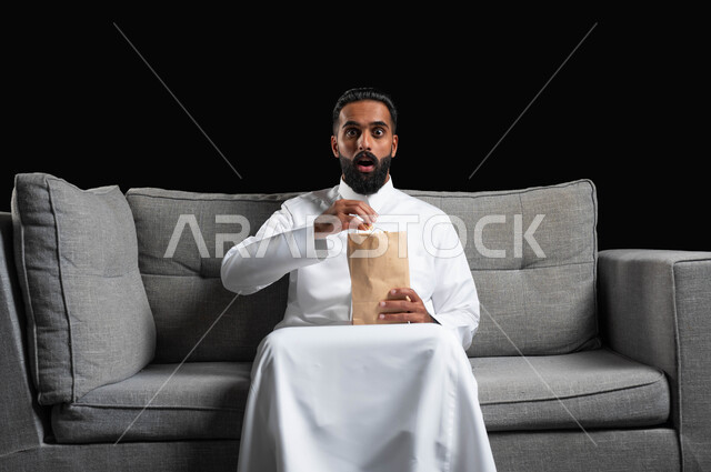 ايماءات وتعابير التعجب والصدمة، الاستمتاع بمشاهدة الأفلام والمسلسلات، بورتريه لرجل عربي خليجي سعودي يرتدي الثوب التقليدي يجلس على اريكة رمادية يقوم بمشاهدة التلفاز وتناول الفوشار،  قضاء أوقات ممتعة ومسلية في المنزل، خلفية سوداء