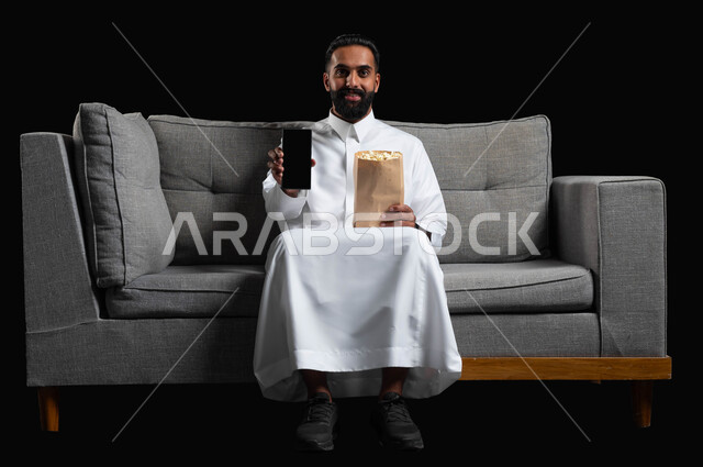 .Gestures of happiness and pleasure, displaying a blank black mobile phone screen, spending enjoyable times watching movies and series, using modern technical devices, a portrait of a smiling Saudi Arabian Gulf man wearing a traditional thobe, sitting on a gray sofa holding a bag of popcorn in his hand, black background.