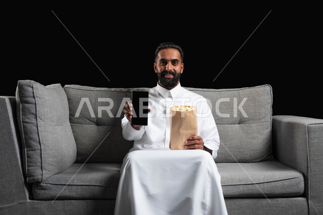 Gestures of happiness and pleasure, displaying a blank black mobile phone screen, spending enjoyable times watching movies and series, using modern technical devices, a portrait of a smiling Saudi Arabian Gulf man wearing a traditional thobe, sitting on a gray sofa holding a bag of popcorn in his hand, black background.