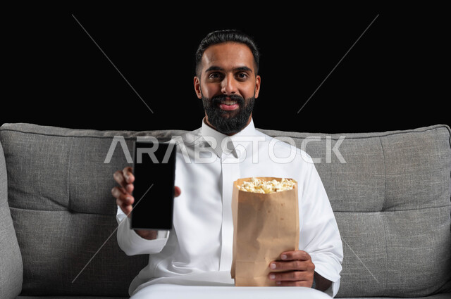 عرض شاشة هاتف محمول سوداء فارغة، قضاء أوقات ممتعة بمشاهدة الأفلام والمسلسلات، استخدام الأجهزة التقنية الحديثة، بورتريه مقرب لشاب عربي سعودي خليجي مبتسم يرتدي الزي التقليدي يجلس على اريكة رمادية يحمل بيده كيس الفوشار، ايماءات السعادة والسرور، خلفية سوداء