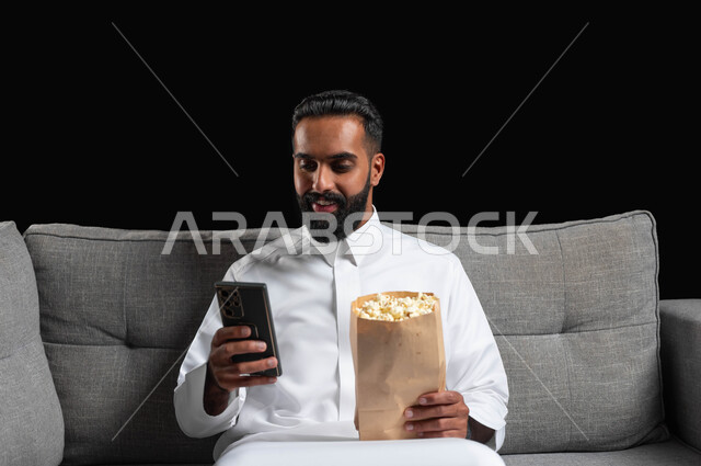 قضاء أوقات ممتعة بمشاهدة الأفلام والمسلسلات، استخدام الأجهزة التكنولوجية الحديثة، بورتريه مقرب لشاب عربي سعودي خليجي مبتسم يرتدي الزي التقليدي يجلس على اريكة رمادية يحمل كيس الفوشار والهاتف المحمول، ايماءات الاندماج والسرور، خلفية سوداء