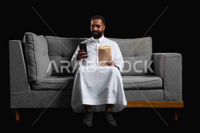 استخدام الأجهزة التكنولوجية الحديثة، بورتريه لشاب عربي سعودي خليجي مبتسم يرتدي الزي التقليدي يجلس على اريكة رمادية يحمل كيس الفوشار والهاتف المحمول، قضاء أوقات ممتعة بمشاهدة الأفلام والمسلسلات، ايماءات الاندماج والسرور، خلفية سوداء