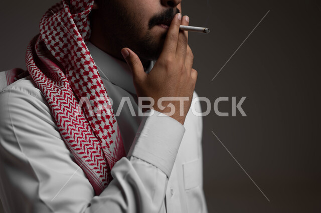 الادمان على التبغ والسجائر، تعاطي مواد ضارة وسامة، عادات سلوكية خاطئة، الإصابه بأمراض الجهاز التنفسي، بورتريه مقرب لشاب عربي سعودي خليجي يرتدي الشماغ والثوب التقليدي يقوم  بسحب نفس عميق من السيجارة، خلفية سوداء