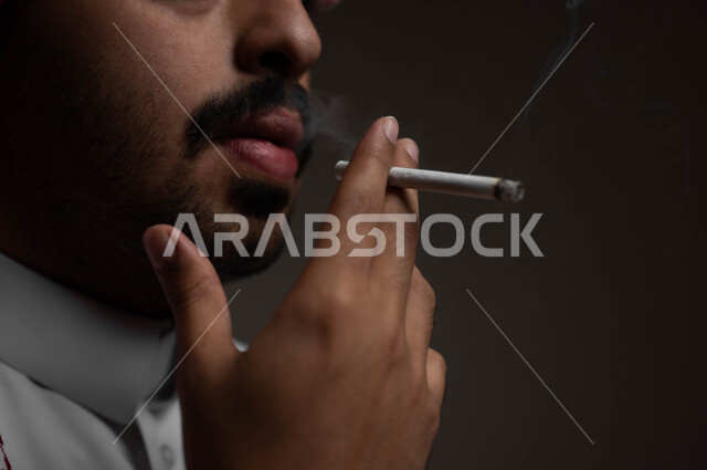 عادات سلوكية خاطئة، الادمان على التبغ والسجائر، تعاطي مواد ضارة وسامة، الإصابه بأمراض الجهاز التنفسي، بورتريه مقرب لشاب عربي سعودي خليجي يرتدي الشماغ والثوب التقليدي يقوم  بسحب نفس عميق من السيجارة، خلفية سوداء