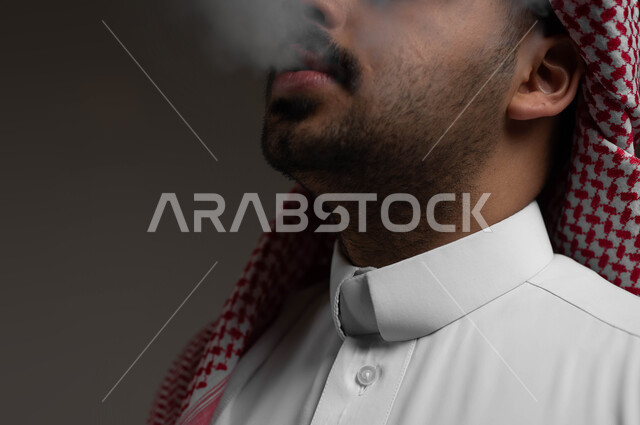 عادات سلوكية خاطئة، الادمان على التبغ والسجائر، تعاطي مواد ضارة وسامة، الإصابه بأمراض الجهاز التنفسي، بورتريه مقرب لشاب عربي سعودي خليجي يرتدي الشماغ والثوب التقليدي يقوم  بسحب نفس عميق من السيجارة، خلفية سوداء