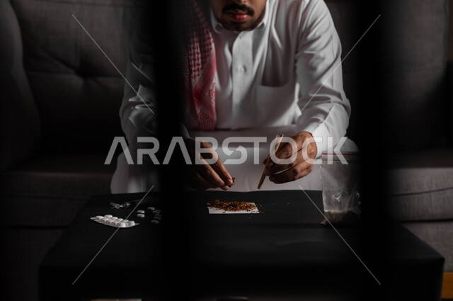 الأثار السلبية للادمان على التبغ والمخدرات، القيام بعادات سلوكية ضارة وتعاطي المواد السامة، المعاناة من الاضطرابات النفسية والمشاكل الصحية، صورة مقربة لرجل عربي خليجي سعودي يرتدي الشماغ والثوب التقليدي يجلس على اريكة يقوم بلف عقب السجائر
