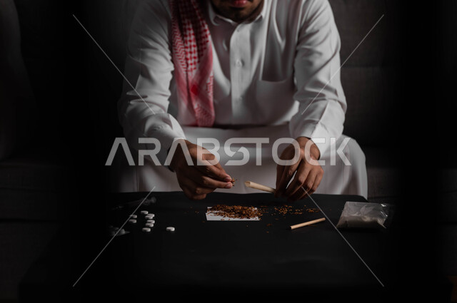 القيام بعادات سلوكية ضارة وتعاطي المواد السامة، المعاناة من الاضطرابات النفسية والمشاكل الصحية، صورة مقربة لرجل عربي خليجي سعودي يرتدي الشماغ والثوب التقليدي يجلس على اريكة يقوم بلف عقب السجائر، الأثار السلبية للادمان على التبغ والمخدرات