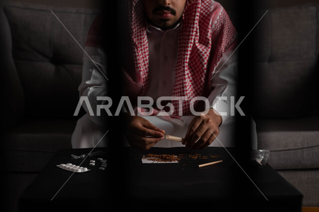 القيام بعادات سلوكية ضارة وتعاطي المواد السامة، المعاناة من الاضطرابات النفسية والمشاكل الصحية، صورة مقربة لرجل عربي خليجي سعودي يرتدي الشماغ والثوب التقليدي يجلس على اريكة يقوم بلف عقب السجائر، الأثار السلبية للادمان على التبغ والمخدرات