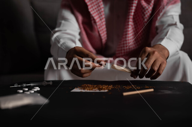 الأثار السلبية للادمان على التبغ والمخدرات، القيام بعادات سلوكية ضارة وتعاطي المواد السامة، المعاناة من الاضطرابات النفسية والمشاكل الصحية، صورة مقربة لرجل عربي خليجي سعودي يرتدي الشماغ والثوب التقليدي يجلس على اريكة يقوم بلف عقب السجائر