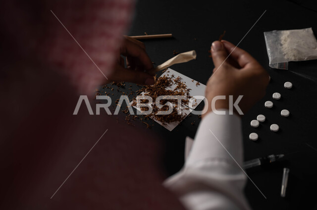 التأثير السلبي للادمان على التبغ والمخدرات، القيام بعادات سلوكية ضارة وتعاطي المواد السامة، المعاناة من الاضطرابات النفسية والمشاكل الصحية، صورة مقربة من الخلف لرجل عربي خليجي سعودي يرتدي الشماغ والثوب التقليدي يجلس على الاريكة يقوم بلف عقب السجائر