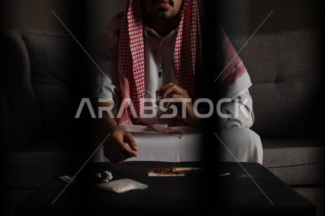 تعاطي المخدرات عن طريق الحقن في الأوردة، الآثار السلبية للادمان، القيام بعادات سلوكية ضارة وتعاطي المواد السامة، المعاناة من الاضطرابات النفسية والمشاكل الصحية، صورة مقربة لرجل عربي خليجي سعودي يرتدي الشماغ والثوب التقليدي يجلس على الاريكة يقوم بأخذ جرعة مخدرة