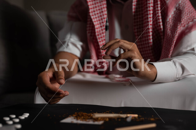 حقن المخدرات في الأوردة، الآثار السلبية للادمان والتعاطي، القيام بعادات سلوكية ضارة وتعاطي المواد السامة، المعاناة من الاضطرابات النفسية والمشاكل الصحية، صورة مقربة لرجل عربي خليجي سعودي يرتدي الشماغ والثوب التقليدي يجلس على الاريكة يقوم بأخذ جرعة مخدرة