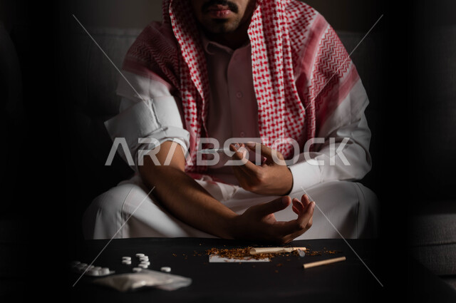 تعاطي المخدرات عن طريق الحقن في الأوردة، الآثار السلبية للادمان، القيام بعادات سلوكية ضارة وتعاطي المواد السامة، المعاناة من الاضطرابات النفسية والمشاكل الصحية، صورة مقربة لرجل عربي خليجي سعودي يرتدي الشماغ والثوب التقليدي يجلس على الاريكة يقوم بأخذ جرعة مخدرة