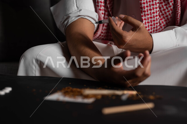 حقن المخدرات في الأوردة، الآثار السلبية للادمان والتعاطي، القيام بعادات سلوكية ضارة وتعاطي المواد السامة، المعاناة من الاضطرابات النفسية والمشاكل الصحية، صورة مقربة لرجل عربي خليجي سعودي يرتدي الشماغ والثوب التقليدي يجلس على الاريكة يقوم بأخذ جرعة مخدرة