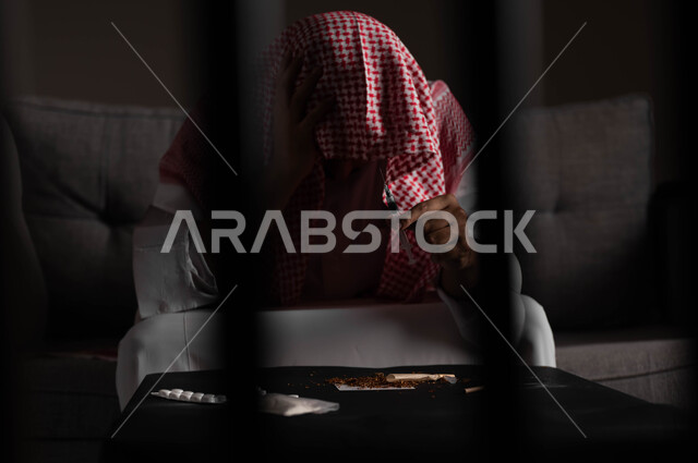 حقن المخدرات في الأوردة، الآثار السلبية للادمان والتعاطي، القيام بعادات سلوكية ضارة وتعاطي المواد السامة، المعاناة من الاضطرابات النفسية والمشاكل الصحية، صورة مقربة لرجل عربي خليجي سعودي يرتدي الشماغ والثوب التقليدي يجلس على الاريكة يقوم بأخذ جرعة مخدرة