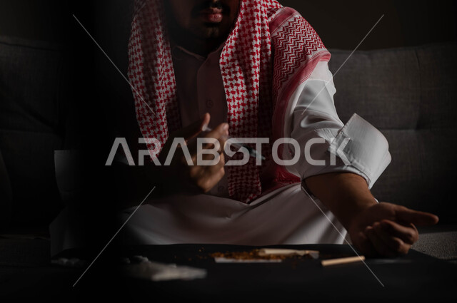 تعاطي المخدرات عن طريق الحقن في الأوردة، الآثار السلبية للادمان، القيام بعادات سلوكية ضارة وتعاطي المواد السامة، المعاناة من الاضطرابات النفسية والمشاكل الصحية، صورة مقربة لرجل عربي خليجي سعودي يرتدي الشماغ والثوب التقليدي يجلس على الاريكة يقوم بأخذ جرعة مخدرة