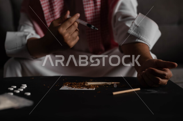 حقن المخدرات في الأوردة، الآثار السلبية للادمان والتعاطي، القيام بعادات سلوكية ضارة وتعاطي المواد السامة، المعاناة من الاضطرابات النفسية والمشاكل الصحية، صورة مقربة لرجل عربي خليجي سعودي يرتدي الشماغ والثوب التقليدي يجلس على الاريكة يقوم بأخذ جرعة مخدرة