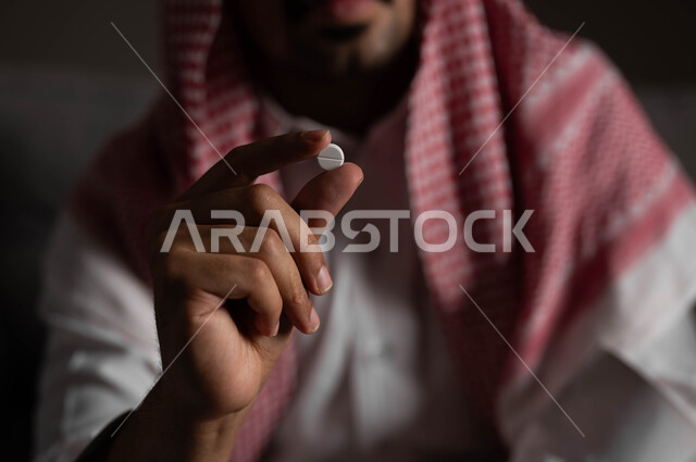 Suffering from psychological disorders and health problems, a portrait of a Saudi Gulf Arab man wearing a shemagh and traditional dress sitting on the sofa holding narcotic pills in his hand, the negative effects of addiction and abuse, engaging in harmful behavioral habits and taking toxic substances