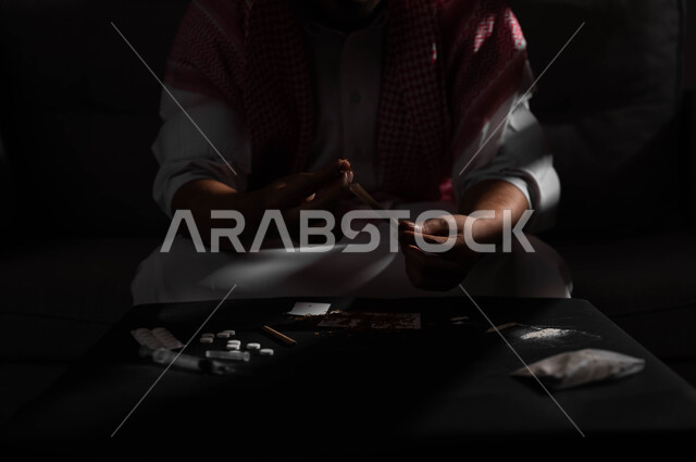 القيام بعادات سلوكية ضارة وتعاطي المواد السامة، المعاناة من الاضطرابات النفسية والمشاكل الصحية، صورة مقربة لرجل عربي خليجي سعودي يرتدي الشماغ والثوب التقليدي يجلس على اريكة يقوم بلف عقب السجائر، الأثار السلبية للادمان على التبغ والمخدرات
