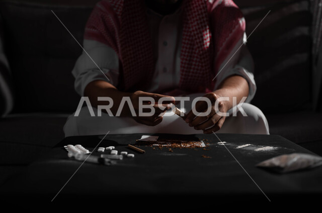 القيام بعادات سلوكية ضارة وتعاطي المواد السامة، المعاناة من الاضطرابات النفسية والمشاكل الصحية، صورة مقربة لرجل عربي خليجي سعودي يرتدي الشماغ والثوب التقليدي يجلس على اريكة يقوم بلف عقب السجائر، الأثار السلبية للادمان على التبغ والمخدرات