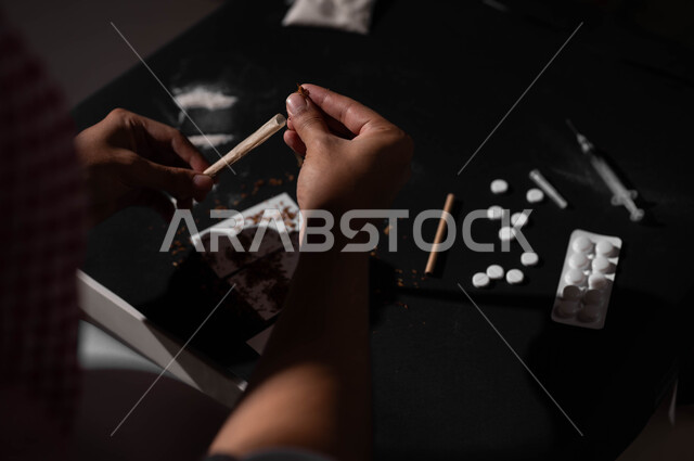 الأثار السلبية للادمان على التبغ والمخدرات، القيام بعادات سلوكية ضارة وتعاطي المواد السامة، المعاناة من الاضطرابات النفسية والمشاكل الصحية، صورة مقربة لرجل عربي خليجي سعودي يرتدي الشماغ والثوب التقليدي يجلس على اريكة يقوم بلف عقب السجائر