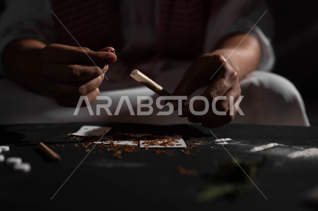 المعاناة من الاضطرابات النفسية والمشاكل الصحية، صورة مقربة لرجل عربي خليجي سعودي يرتدي الشماغ والثوب التقليدي يجلس على اريكة يقوم بلف عقب السجائر، الأثار السلبية للادمان على التبغ والمخدرات، القيام بعادات سلوكية ضارة وتعاطي المواد السامة