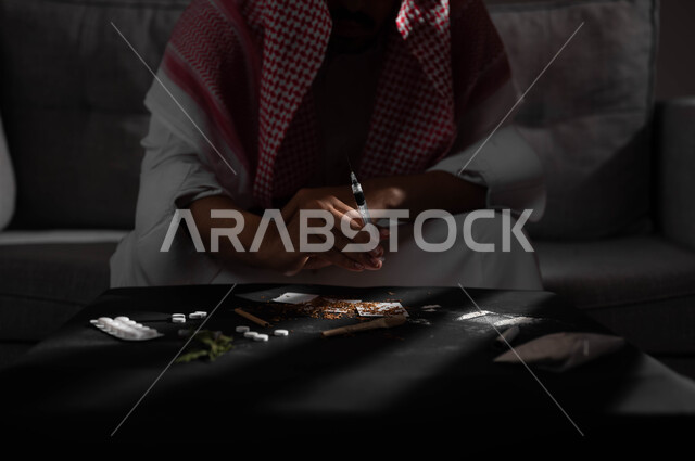 المعاناة من الاضطرابات النفسية والمشاكل الصحية، حقن المخدرات في الأوردة، الآثار السلبية للادمان، القيام بعادات سلوكية ضارة وتعاطي المواد السامة، صورة مقربة لرجل عربي خليجي سعودي يرتدي الشماغ والثوب التقليدي يجلس على الاريكة يقوم بأخذ ابرة مخدرة