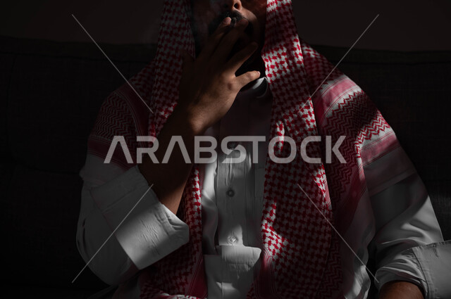 الادمان على التبغ والسجائر، تعاطي مواد ضارة وسامة، عادات سلوكية خاطئة، الإصابه بأمراض الجهاز التنفسي، بورتريه مقرب لشاب عربي سعودي خليجي يرتدي الشماغ والثوب التقليدي يقوم  بسحب نفس عميق من السيجارة، خلفية سوداء