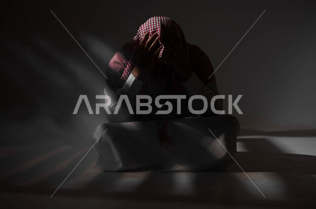 الإنعزال النفسي والشخصية الانطوائية، معاقبة النفس وتأنيب الضمير، الآثار السلبية للادمان والتعاطي، المعاناة من الاضطرابات النفسية، بورتريه لرجل عربي خليجي سعودي يرتدي الزي التقليدي يجلس على الارض مصاب بالإكتئاب والإنهيار العصبي، خلفية سوداء