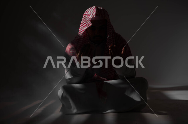 إيماءات تدل على التوسل والتضرع، مفهوم الدعاء والترجي والطلب، بورتريه لشاب عربي خليجي سعودي يرتدي الشماغ والثوب التقليدي يجلس على الأرض يرفع يديه ويدعو الله، التوبة والرجوع الى الله، الاجتهاد في العباده والطاعة في شهر رمضان المبارك, خلفية سوداء