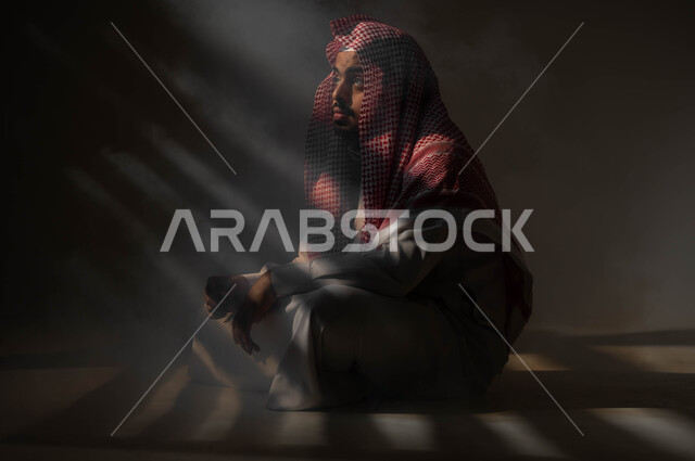 الشعور بالملل والتعب من ضغوطات الحياة والعمل، بورتريه لرجل عربي خليجي سعودي يرتدي الثوب والشماغ التقليدي يجلس على الارض ينظر إلى شيء ما بإيماءات التفكير، التمعن والتركيز وشرود الذهن، التأمل والتخطيط للمستقبل، إيجاد حل لمشكلة ما، خلفية سوداء