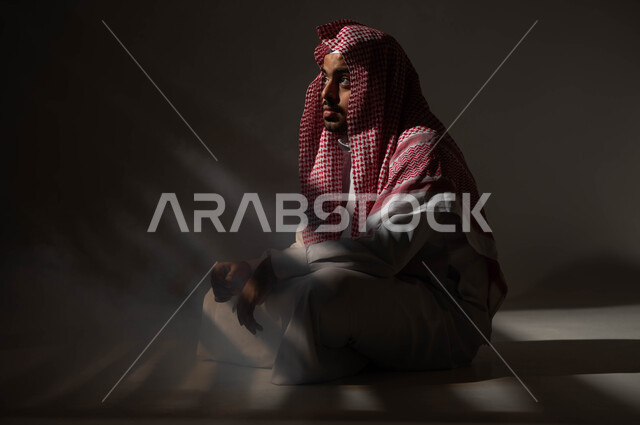 Meditation and planning for the future, finding a solution to a problem, feeling bored and tired from the pressures of life and work, a portrait of a Saudi Gulf Arab man wearing the traditional thobe and shemagh, sitting on the ground looking at something with gestures of thought, contemplation, concentration and absent-mindedness, a black background.