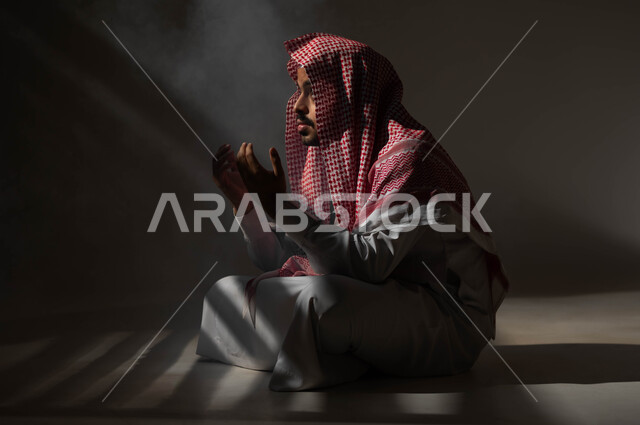 إيماءات تدل على التوسل والتضرع، مفهوم الدعاء والترجي والطلب، بورتريه لشاب عربي خليجي سعودي يرتدي الشماغ والثوب التقليدي يجلس على الأرض يرفع يديه ويدعو الله، التوبة والرجوع الى الله، الاجتهاد في العباده والطاعة في شهر رمضان المبارك, خلفية سوداء