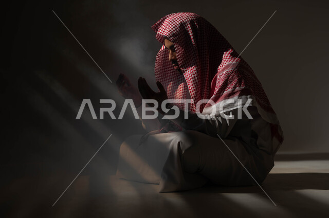 تعابير تدل على التوسل والتضرع، مفهوم الدعاء والترجي والطلب، بورتريه لشاب عربي خليجي سعودي يرتدي الشماغ والثوب التقليدي يجلس على الأرض يرفع يديه ويدعو الله، التوبة والرجوع الى الله، الاجتهاد في العباده والطاعة في شهر رمضان المبارك, خلفية سوداء