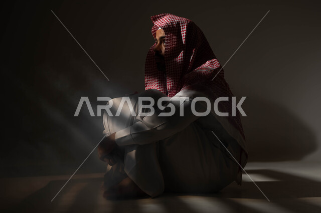 معاقبة النفس وتأنيب الضمير، الآثار السلبية للادمان والتعاطي، المعاناة من الاضطرابات النفسية، الإنعزال النفسي والشخصية الانطوائية، بورتريه لرجل عربي خليجي سعودي يرتدي الزي التقليدي يجلس على الارض مصاب بالإكتئاب والإنهيار العصبي، خلفية سوداء