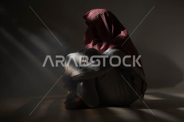 الآثار السلبية للادمان والتعاطي، معاقبة النفس وتأنيب الضمير، المعاناة من الاضطرابات النفسية، الإنعزال النفسي والشخصية الانطوائية، بورتريه لرجل عربي خليجي سعودي يرتدي الزي التقليدي يجلس على الارض مصاب بالإكتئاب والإنهيار العصبي، خلفية سوداء
