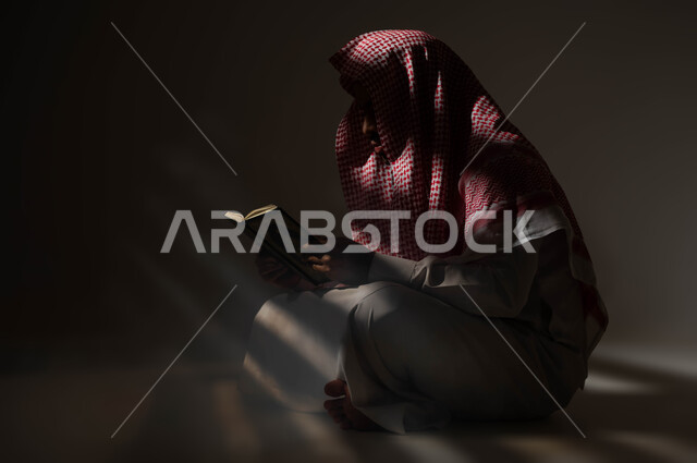 الإكثار من الذكر والإجتهاد في العباده في شهر رمضان، الشعور بالخشوع والطمأنينه, تلاوة وقراءة القرآن الكريم، التوبة والرجوع الى الله تعالى، بورتريه مقرب لرجل عربي سعودي خليجي يرتدي الشماغ والثوب التقليدي يحمل بيده المصحف الشريف, خلفية سوداء
