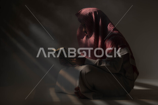 الشعور بالخشوع والطمأنينه, الإكثار من الذكر والإجتهاد في العباده في شهر رمضان، تلاوة وقراءة القرآن الكريم، التوبة والرجوع الى الله تعالى، بورتريه مقرب لرجل عربي سعودي خليجي يرتدي الشماغ والثوب التقليدي يحمل بيده المصحف الشريف, خلفية سوداء