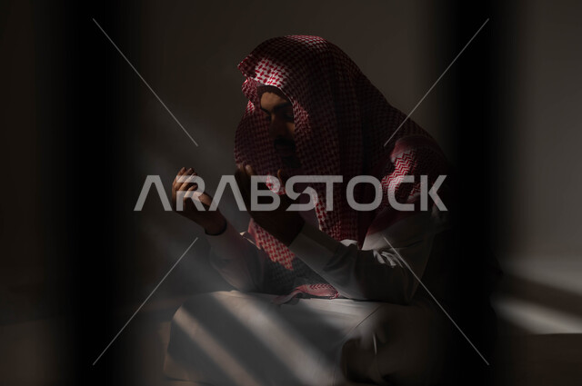 إيماءات تدل على التوسل والتضرع، مفهوم الدعاء والترجي والطلب، بورتريه لشاب عربي خليجي سعودي يرتدي الشماغ والثوب التقليدي يجلس على الأرض يرفع يديه ويدعو الله، التوبة والرجوع الى الله، الاجتهاد في العباده والطاعة في شهر رمضان المبارك, خلفية سوداء
