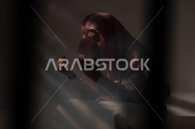 تعابير تدل على التوسل والتضرع، مفهوم الدعاء والترجي والطلب، بورتريه لشاب عربي خليجي سعودي يرتدي الشماغ والثوب التقليدي يجلس على الأرض يرفع يديه ويدعو الله، التوبة والرجوع الى الله، الاجتهاد في العباده والطاعة في شهر رمضان المبارك, خلفية سوداء