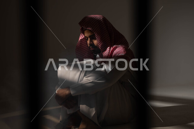 Suffering from psychological disorders, a portrait of a Saudi Gulf Arab man wearing traditional clothing sitting on the ground with expressions of psychological isolation and introverted personality, negative effects of addiction and abuse, depression and nervous breakdown, self-punishment and remorse, black background.