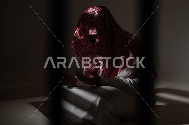 The concept of supplication, begging, and request, a portrait of a Saudi Gulf Arab young man wearing a shemagh and traditional dress, sitting on the ground, raising his hands and calling upon God with gestures that indicate pleading and supplication, repentance and returning to God, diligence in worship and obedience in the blessed month of Ramadan, black background.