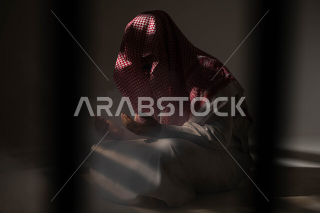 الاجتهاد في العباده والطاعة في شهر رمضان المبارك, مفهوم الدعاء والترجي والطلب، بورتريه لشاب عربي خليجي سعودي يرتدي الشماغ والثوب التقليدي يجلس على الأرض يرفع يديه ويدعو الله بإيماءات تدل على التوسل والتضرع، التوبة والرجوع الى الله، خلفية سوداء