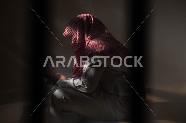 التوبة والرجوع الى الله، الاجتهاد في العباده والطاعة في شهر رمضان المبارك, مفهوم الدعاء والترجي والطلب، بورتريه لشاب عربي خليجي سعودي يرتدي الشماغ والثوب التقليدي يجلس على الأرض يرفع يديه ويدعو الله بإيماءات تدل على التوسل والتضرع، خلفية سوداء