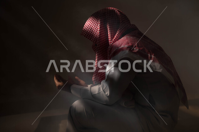 مفهوم الدعاء والترجي والطلب، بورتريه لشاب عربي خليجي سعودي يرتدي الشماغ والثوب التقليدي يجلس على الأرض يرفع يديه ويدعو الله بإيماءات تدل على التوسل والتضرع، التوبة والرجوع الى الله، الاجتهاد في العباده والطاعة في شهر رمضان المبارك, خلفية سوداء